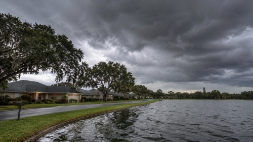 Established lakefront homes with architectural shingle roofs and mature live oak trees under dark tropical storm clouds in Auburndale Florida.