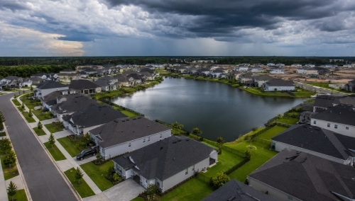 Aerial drone view of a modern master-planned residential subdivision in Haines City Florida showing homes with dark roofs surrounding a community retention pond under dramatic storm clouds.