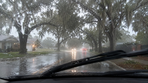 A driver's perspective through a rain-streaked windshield with active wipers on a wet residential street lined with large oak trees and Spanish moss in Lake Wales Florida.